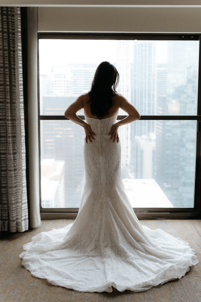 Bride standing with hands on her hips facing a high rise window view of city buildings.