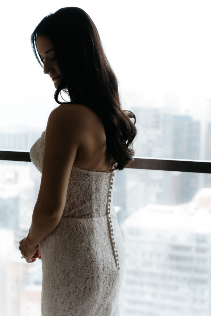 Bride standing by a tall window showing the back details of her lace strapless dress.