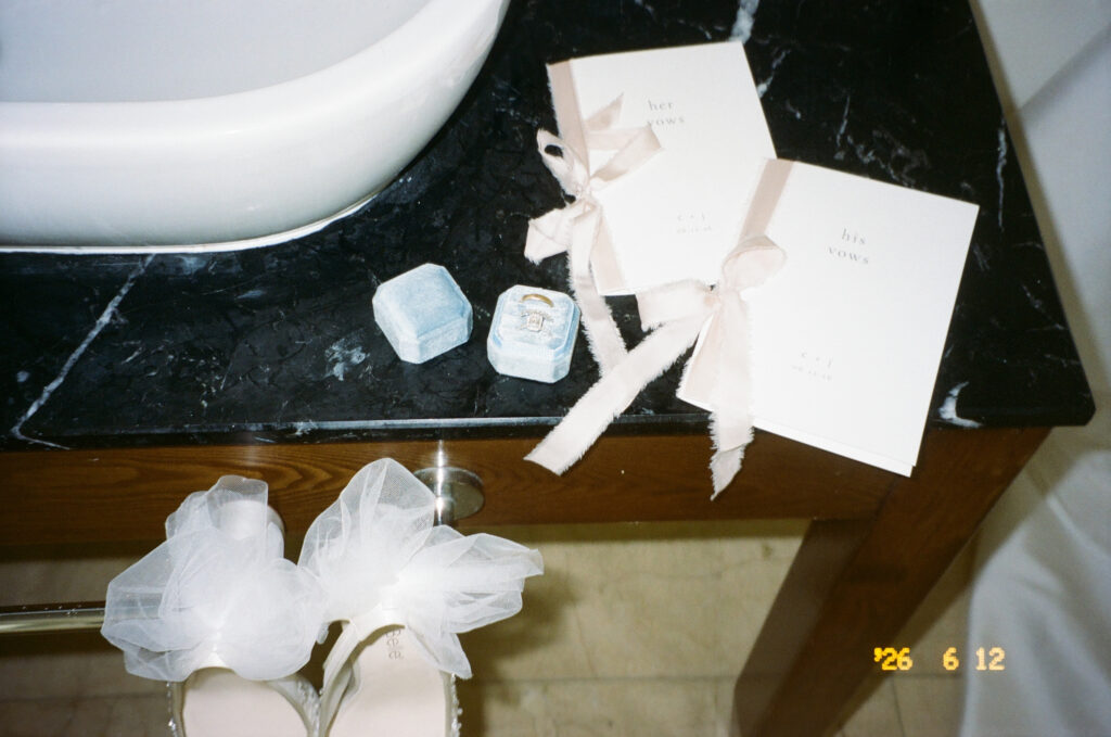 Vow books, ring boxes, and bridal shoes arranged on a dark marble counter captured on film.