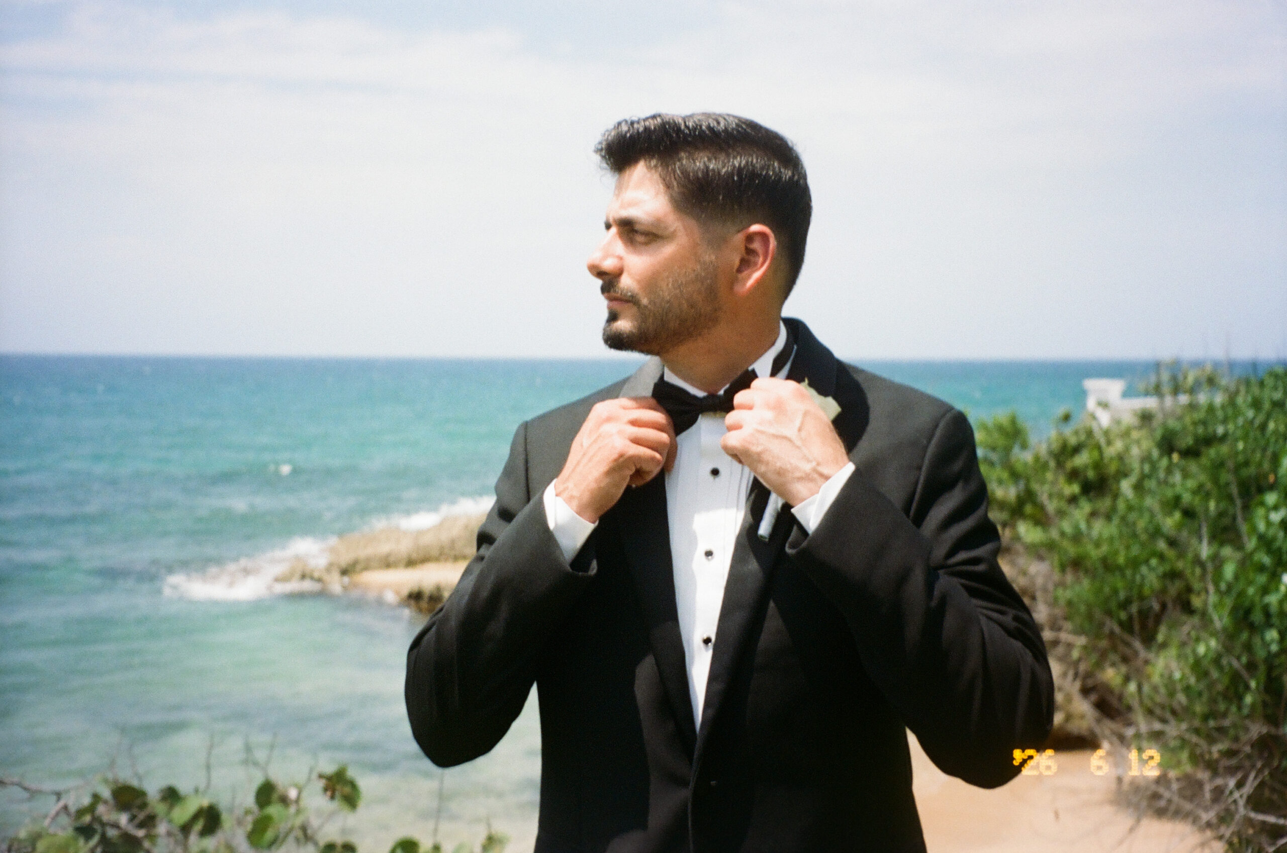 Groom in a tuxedo adjusting his bowtie on a sunny coast with ocean waves behind him.