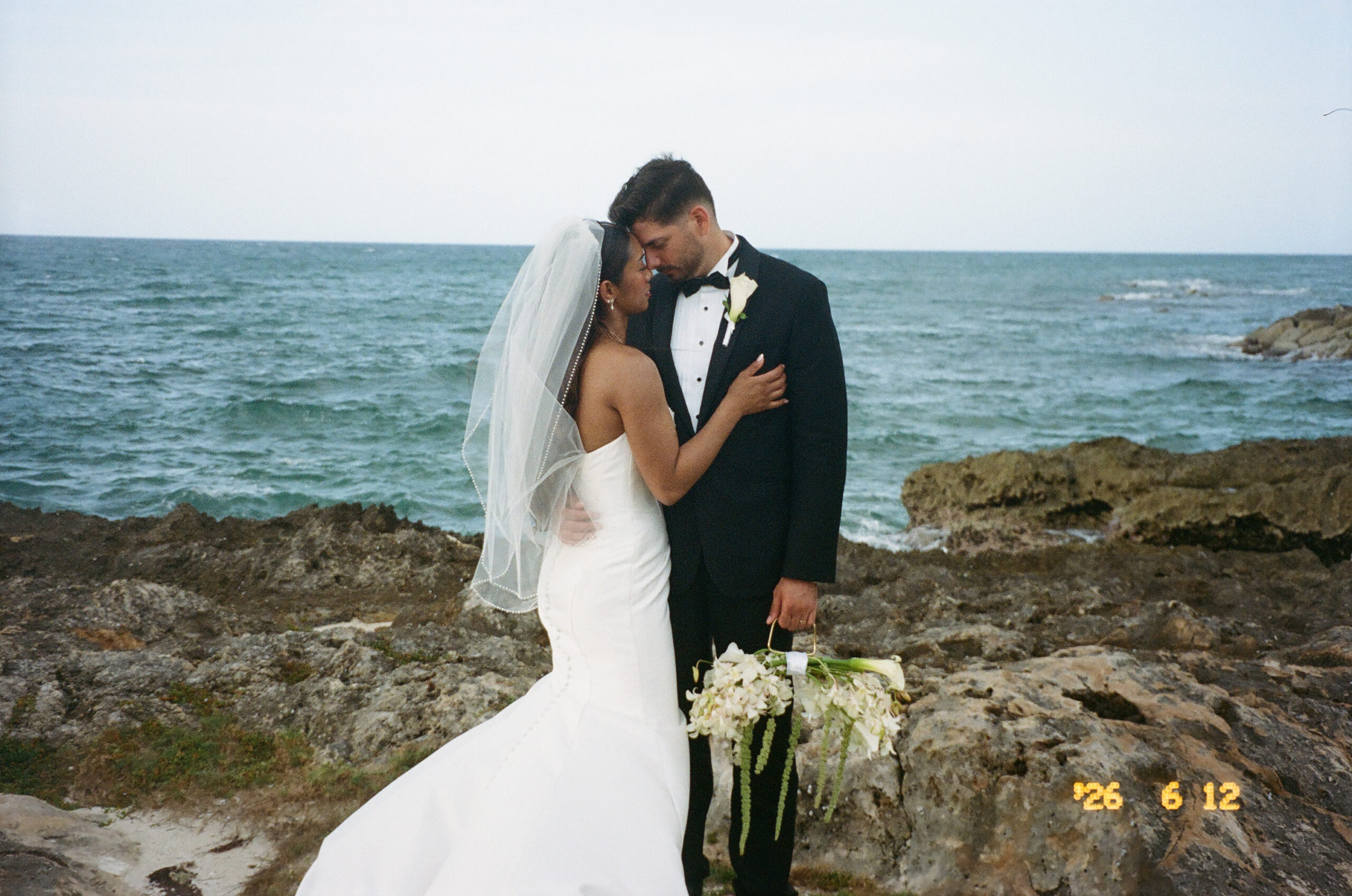 Bride and groom touching foreheads while standing on rocky ocean shores captured on 35mm film.